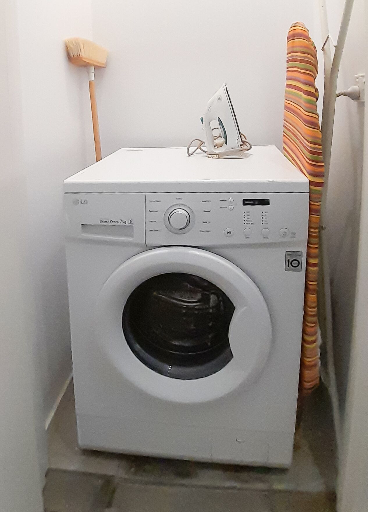 A compact, white front-loading washing machine in a small laundry room with a broom and striped curtain.