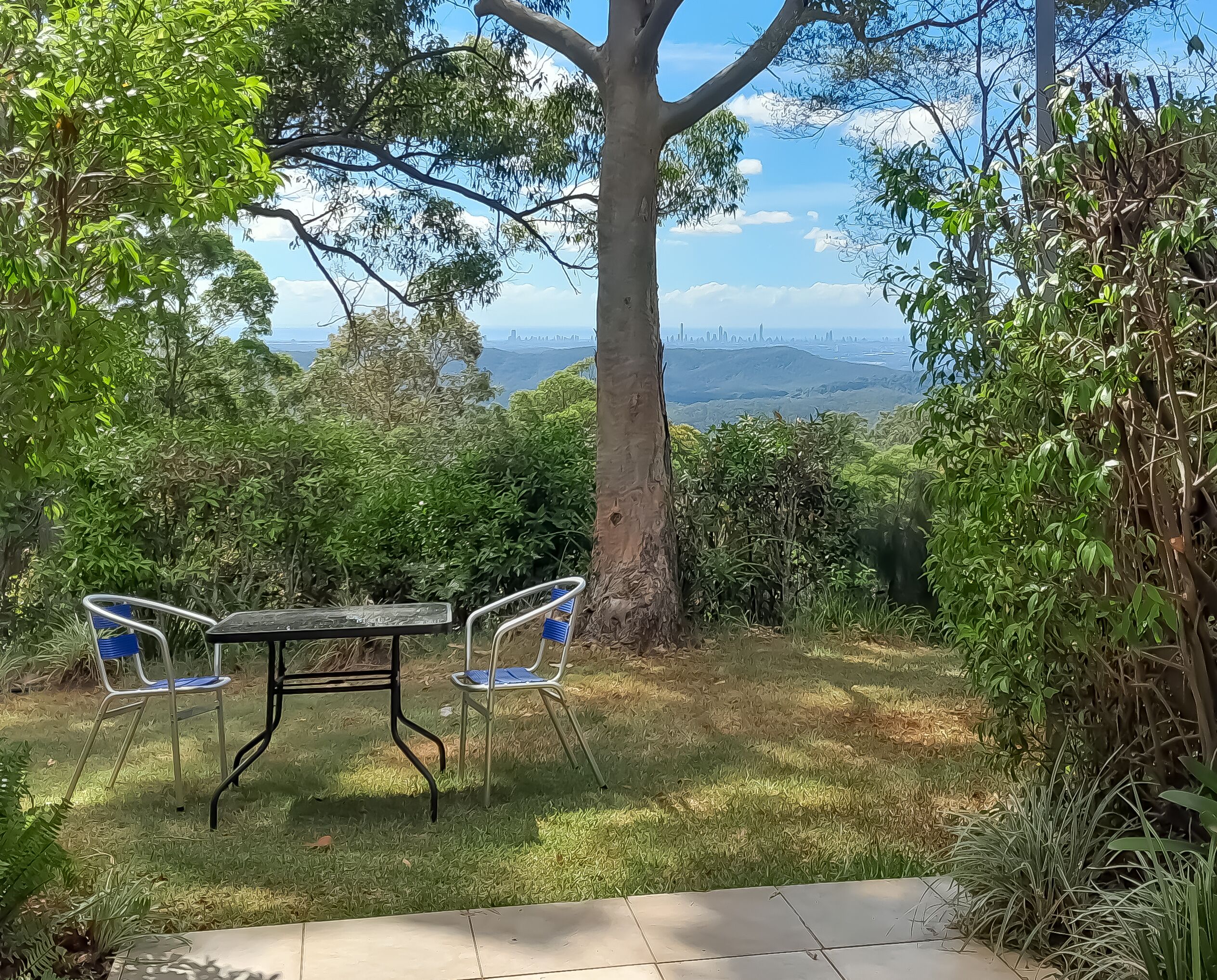 Un jardín con una mesa y sillas, rodeado de árboles y vistas a un paisaje urbano lejano.