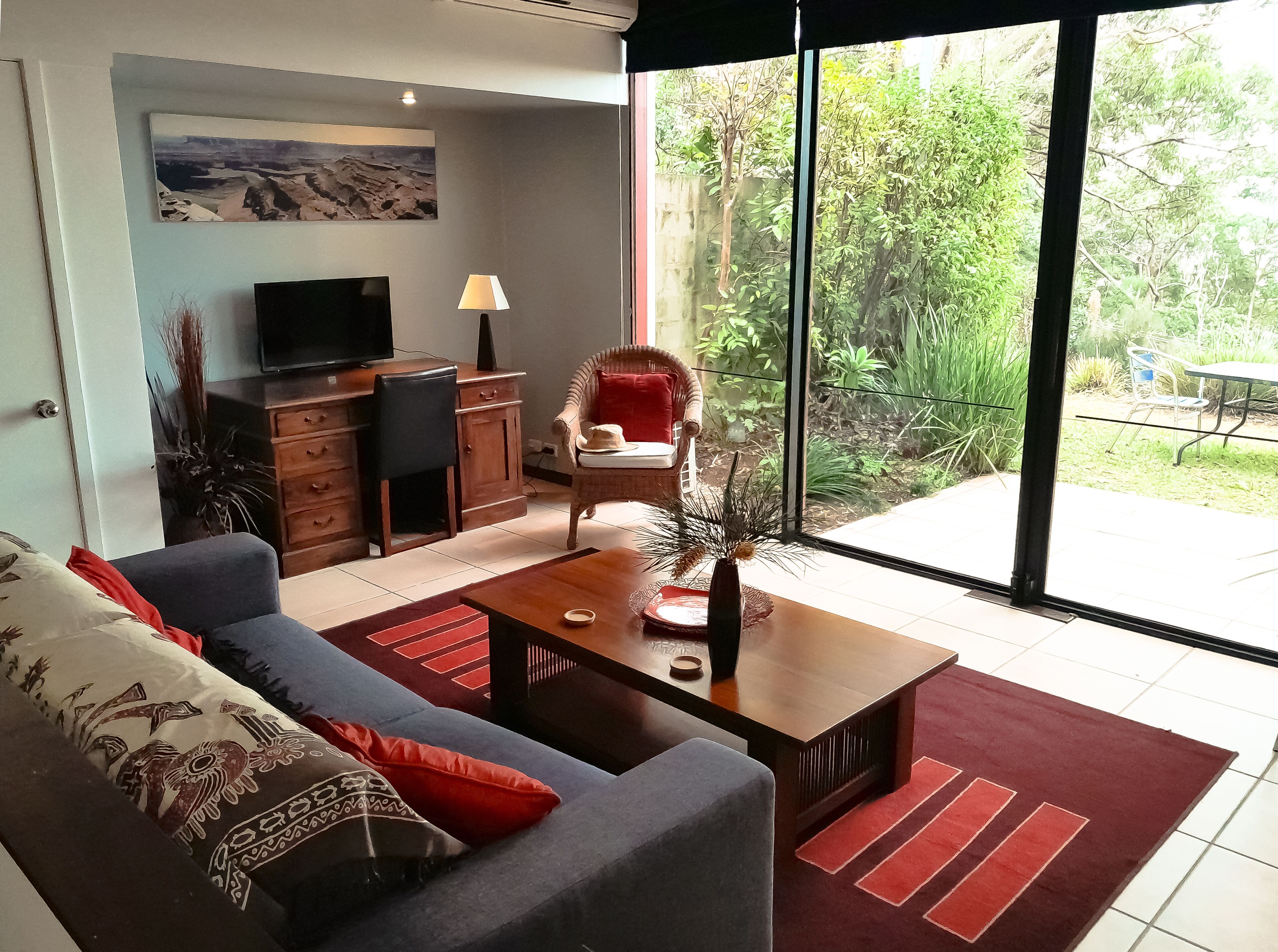 Un salon moderne avec un canapé, une table basse, un téléviseur et une vue sur un jardin grâce à de grandes fenêtres.