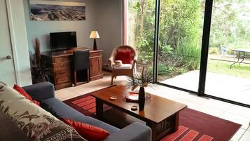 Un salon moderne avec un canapé, une table basse, un téléviseur et une vue sur un jardin grâce à de grandes fenêtres.