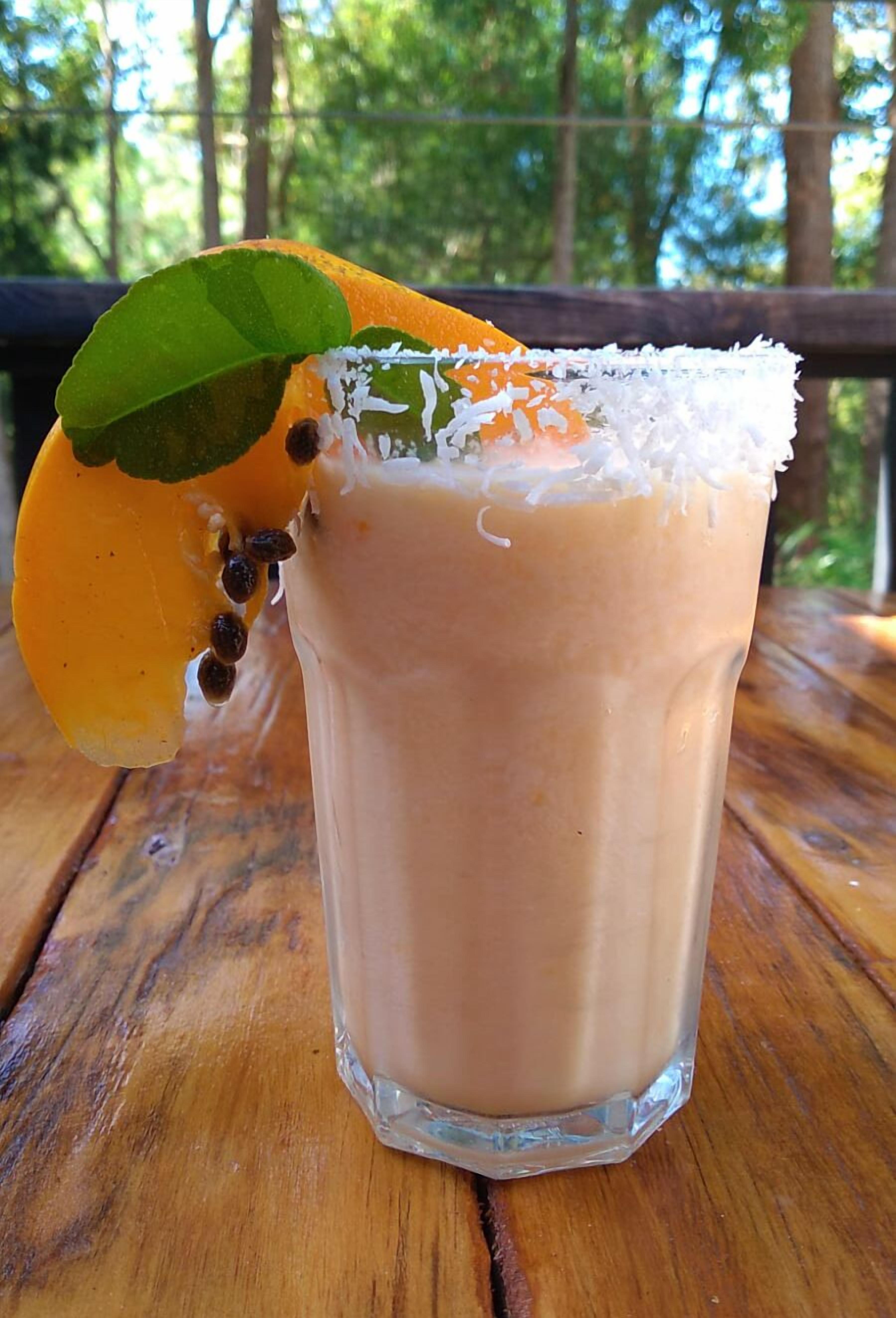 A glass of beverage with a slice of fruit and coconut on the rim, placed on a wooden surface.