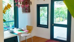 A room with a wooden ceiling, a desk with a chair, a window with a view of greenery, and a door with a mat. - Sky Island Studios (Clagiraba)