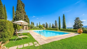 Una piscina con una tumbona y sombrilla, rodeada de césped cuidado y altos árboles.