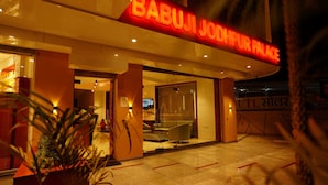 The exterior of Babuji Johpur Palace hotel with a prominent sign, a glass entrance, and a well-lit interior visible through the doors.