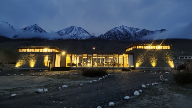 A building with a glass facade and a flag, surrounded by snow-covered mountains and a cloudy sky. - The Merak (Leh)