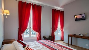 A hotel room with a bed, two windows with red curtains, a small table with a lamp, and a TV on the wall. - Logis Hotel de la Tour (Libourne)