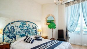 A bedroom with a large bed, a headboard with a blue landscape painting, a small table with a lamp, a floor lamp, and a view of the outdoors through a window with curtains. - Logis Hotel de la Tour (Libourne)