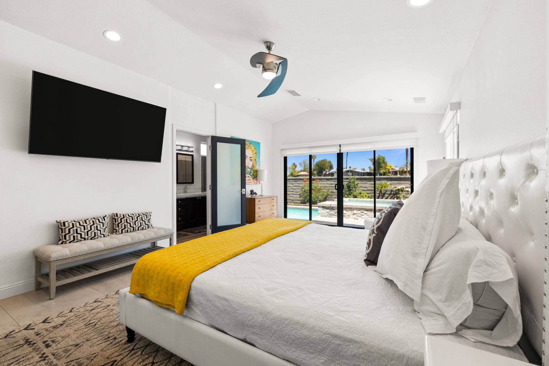 A modern bedroom with a large bed, a flat-screen TV, a ceiling fan, and a view of a pool.