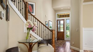 A hallway with a wooden staircase, a glass table with a vase of flowers, a chair, and a door leading to a bright room. - Impeccable Décor and Furnishing, Splendid Neighborhood, Pet Friendly, Fenced Backyard, Next to Park (Portland)