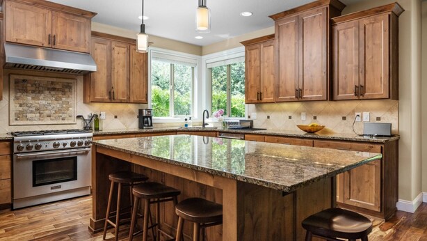 A spacious kitchen with wooden cabinets, a large island with stools, a gas stove, and a window providing a view of greenery. - Impeccable Décor and Furnishing, Splendid Neighborhood, Pet Friendly, Fenced Backyard, Next to Park (Portland)