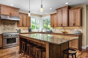 A spacious kitchen with wooden cabinets, a large island with stools, a gas stove, and a window providing a view of greenery. - Impeccable Décor and Furnishing, Splendid Neighborhood, Pet Friendly, Fenced Backyard, Next to Park (Portland)