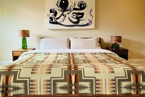 A bed with a patterned blanket, two bedside tables with lamps, and a wall with a large abstract artwork. - Impeccable Décor and Furnishing, Splendid Neighborhood, Pet Friendly, Fenced Backyard, Next to Park (Portland)