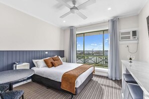 A hotel room with a bed, a desk, a chair, a small table, a ceiling fan, and a large window with a city view.