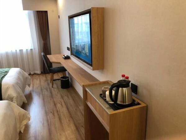 A hotel room with a wooden desk, a television mounted on the wall, a bed, and a small kitchenette area with a kettle and cups.