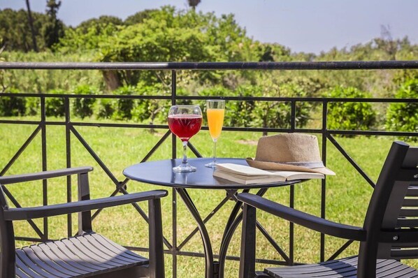 A table with two glasses of red and orange beverages, a book, and a straw hat on a balcony with a metal railing. - Astra Village Suites & Spa, Kefalonia (Kefalonia)