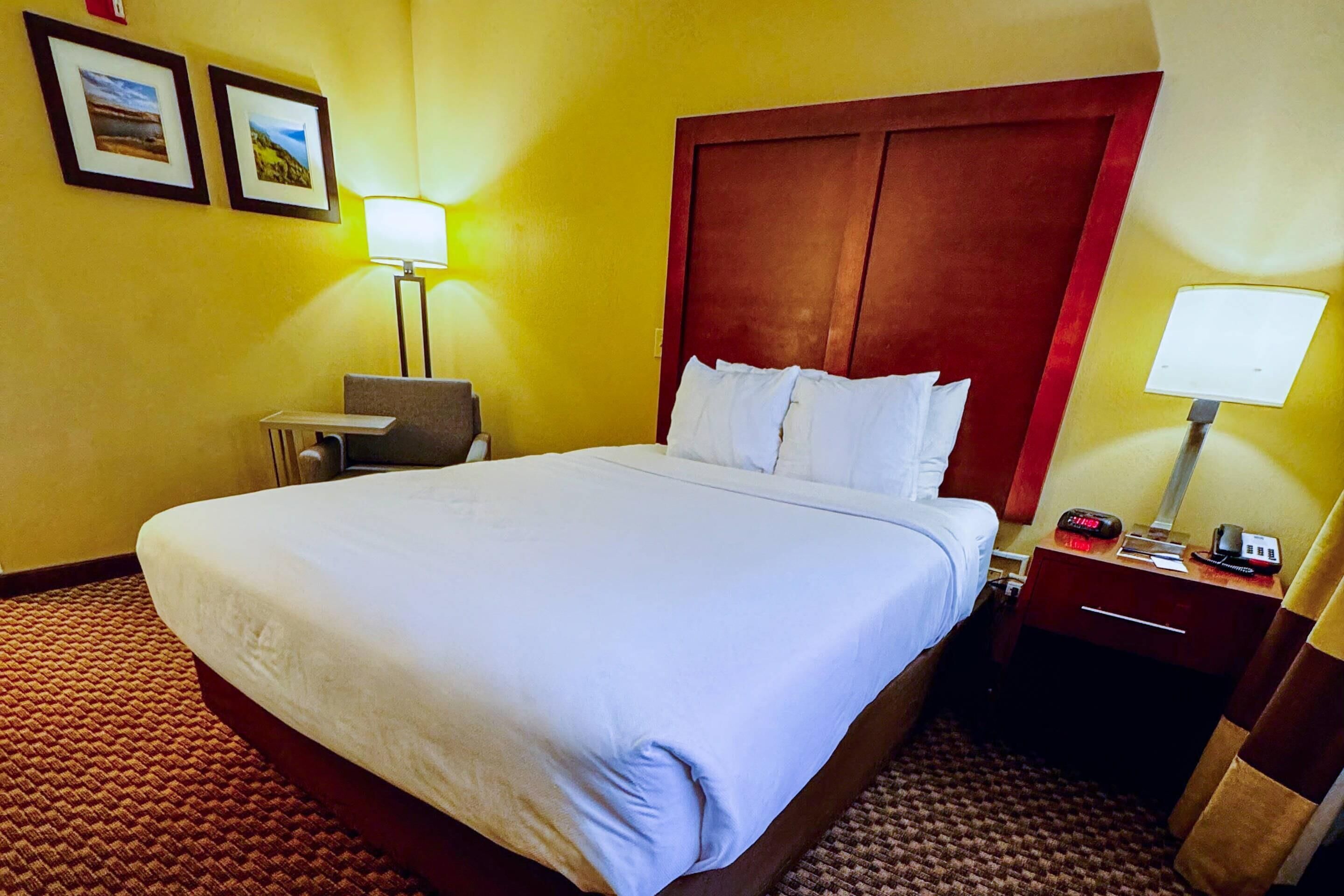 A hotel room with a large bed, a desk with a chair, a nightstand with a lamp, and two framed pictures on the wall.