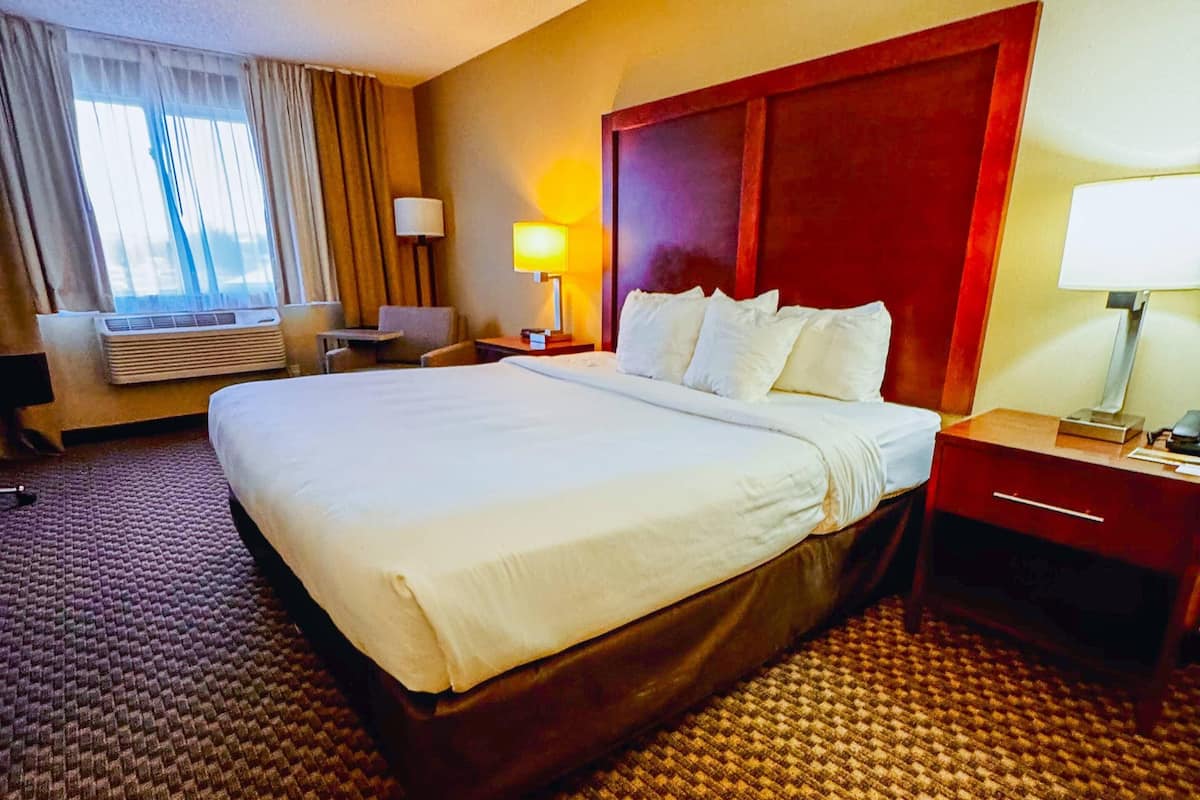 A hotel room with a large bed, a desk, a chair, an air conditioning unit, and a window with curtains.