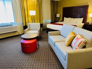A hotel room with a bed, a sofa, a chair, a small table, a red ottoman, and a carpeted floor.