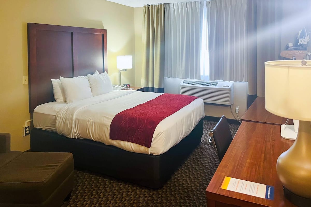 A hotel room with a bed, bedside table, lamp, air conditioning unit, and a desk with a chair.