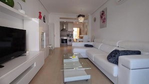 A modern living room with a flat-screen TV, a glass coffee table, a white sofa, and a kitchen in the background. - stunning apartment overlooking the pool (Orihuela)
