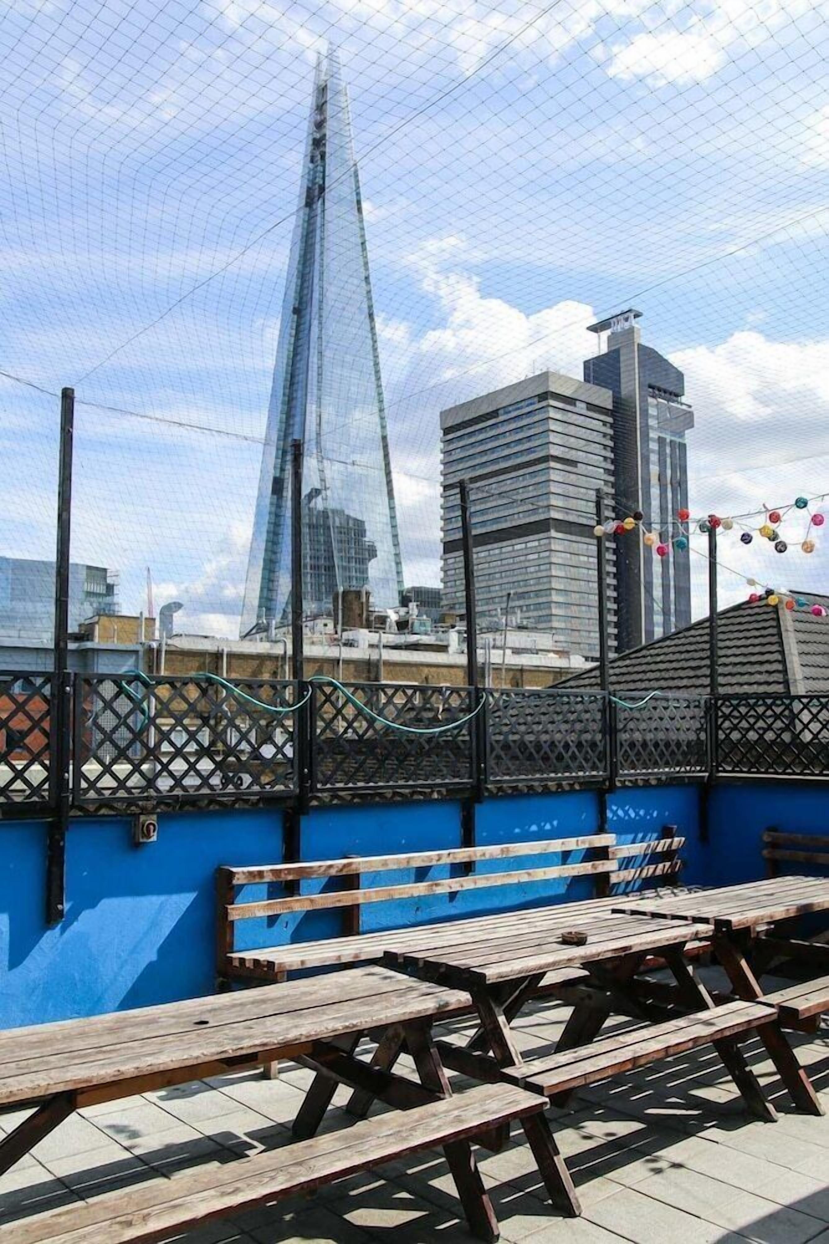 Une terrasse sur le toit, avec des tables de pique-nique en bois et un mur bleu, offrant une vue sur un paysage urbain dominé par des bâtiments modernes.
