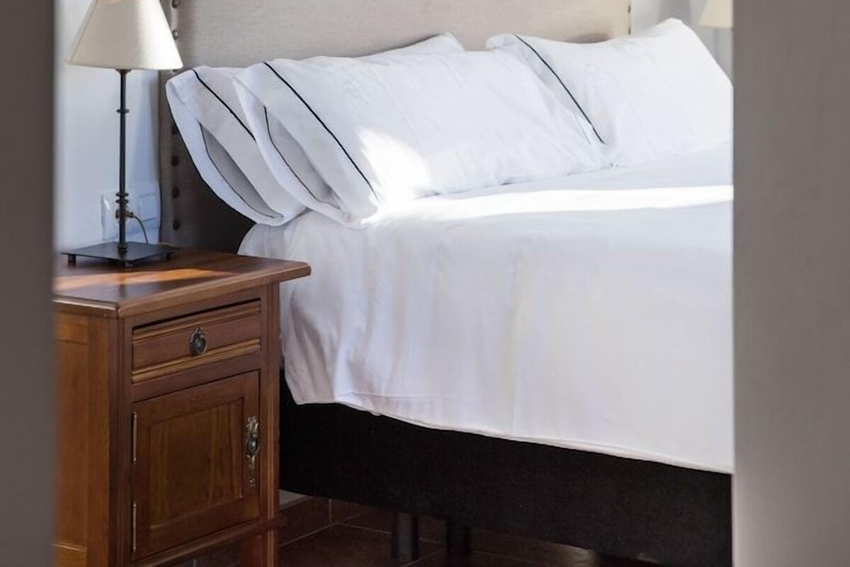 Una cama bien hecha con cabecera, lámpara de noche y mesita de madera.