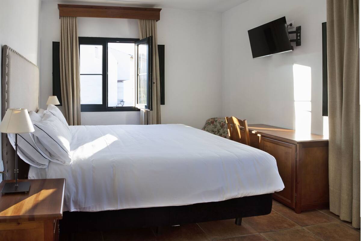 Une chambre d’hôtel avec un lit, un téléviseur fixé au mur, un bureau avec une lampe et une fenêtre avec des rideaux.