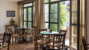 A dining area with multiple tables and chairs, large windows with curtains, and a potted plant on a small table.