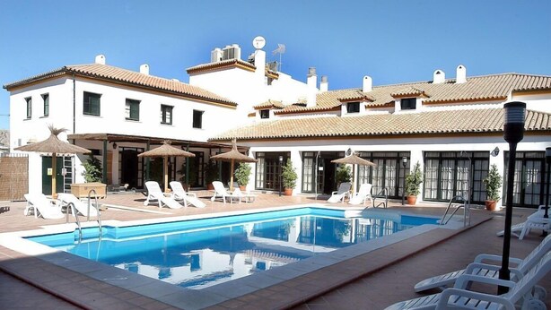 A swimming pool with lounge chairs and umbrellas, surrounded by a building with multiple windows and a tiled roof.