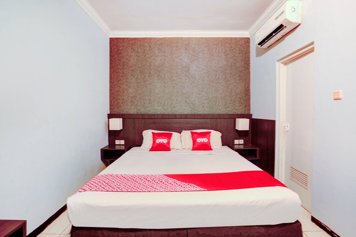 Quarto de hotel com uma cama, colcha vermelha e branca, duas almofadas com a marca "OYO", cabeceira de madeira, ar condicionado fixado na parede e uma porta à direita.