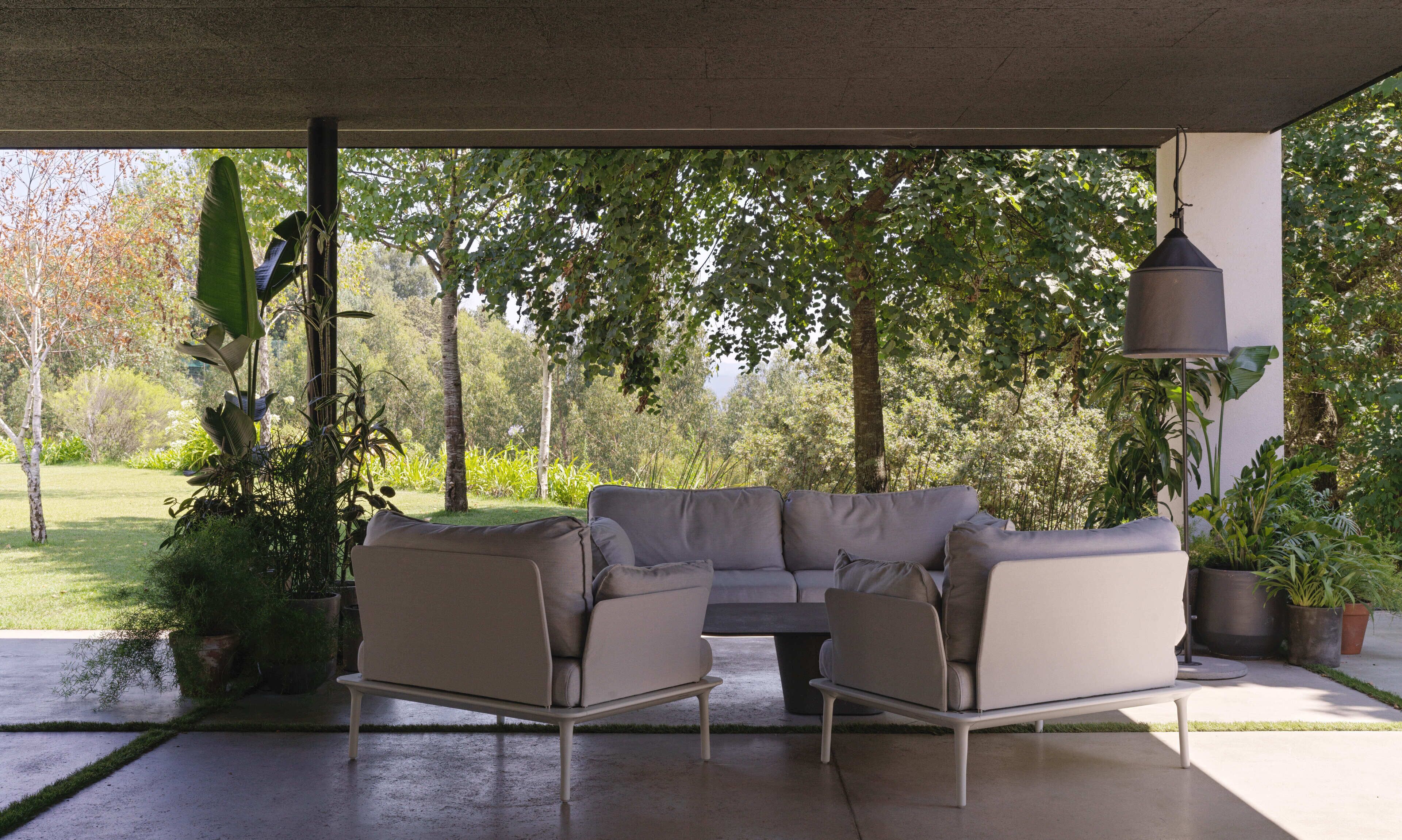 A covered outdoor seating area with a modern sofa and table, surrounded by lush greenery and trees.