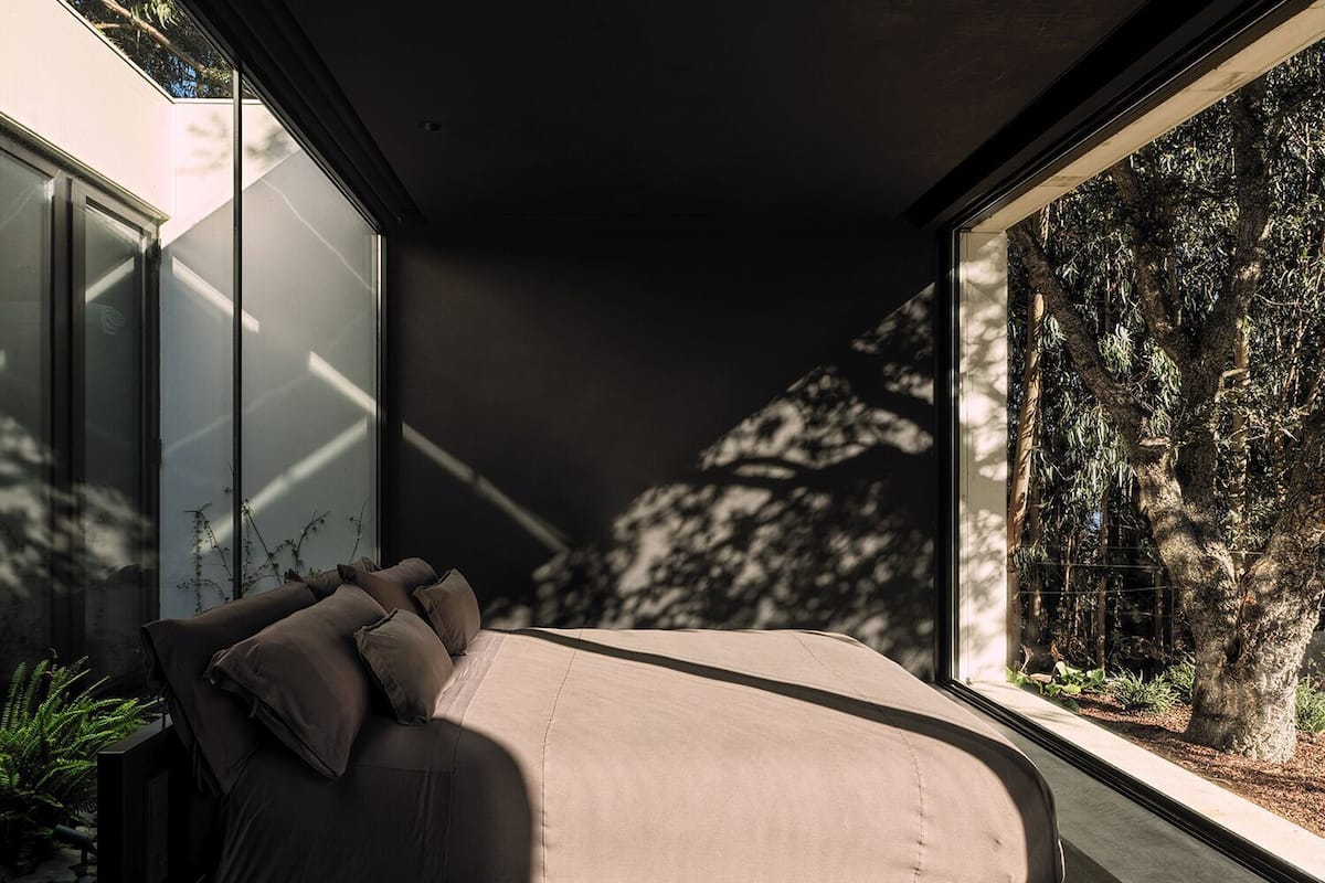 A modern bedroom with a bed, large windows, and a view of trees.