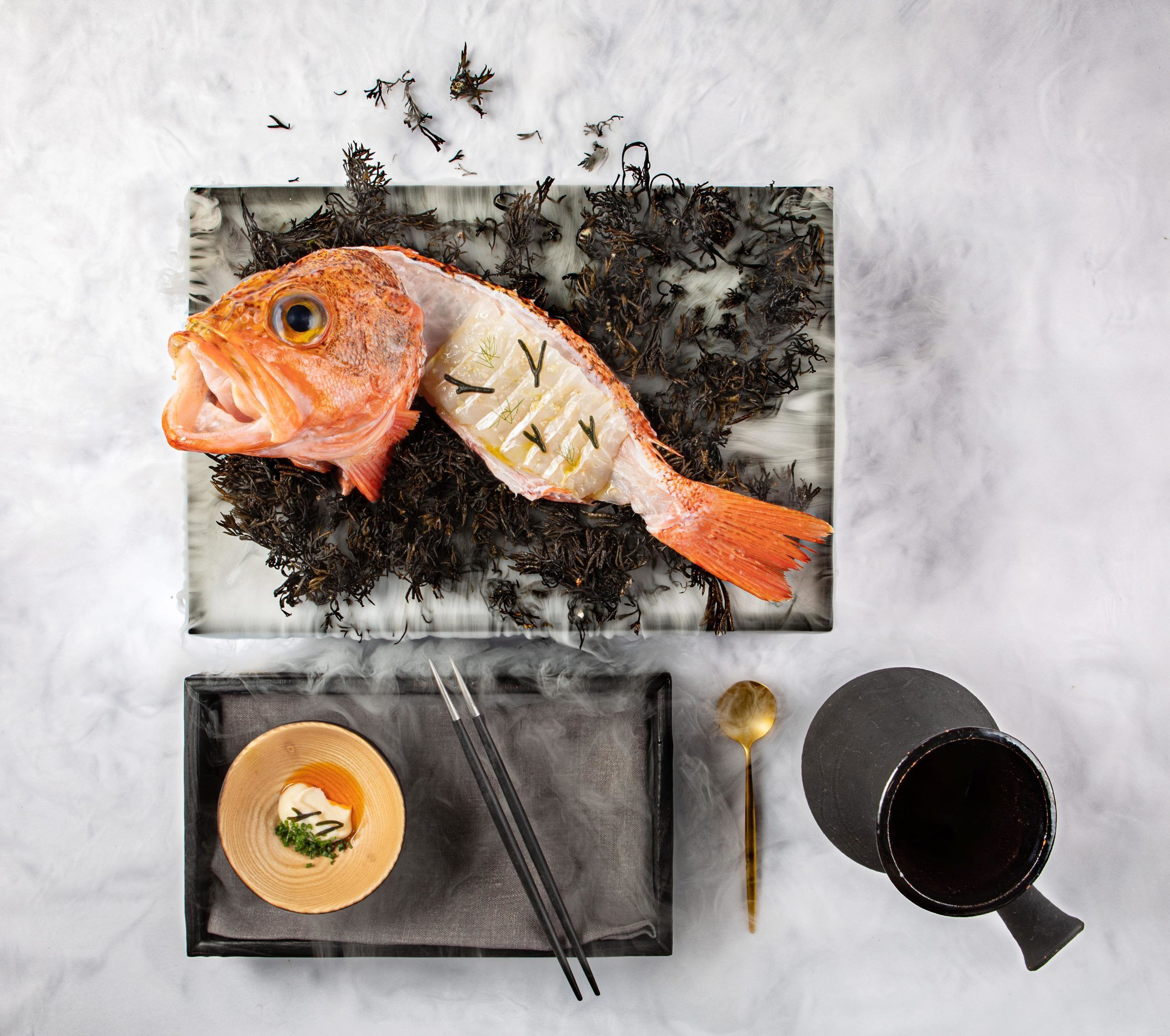 Vista cenital de una mesa de comedor con pescado, algas marinas y un bol de sopa sobre una superficie de mármol.