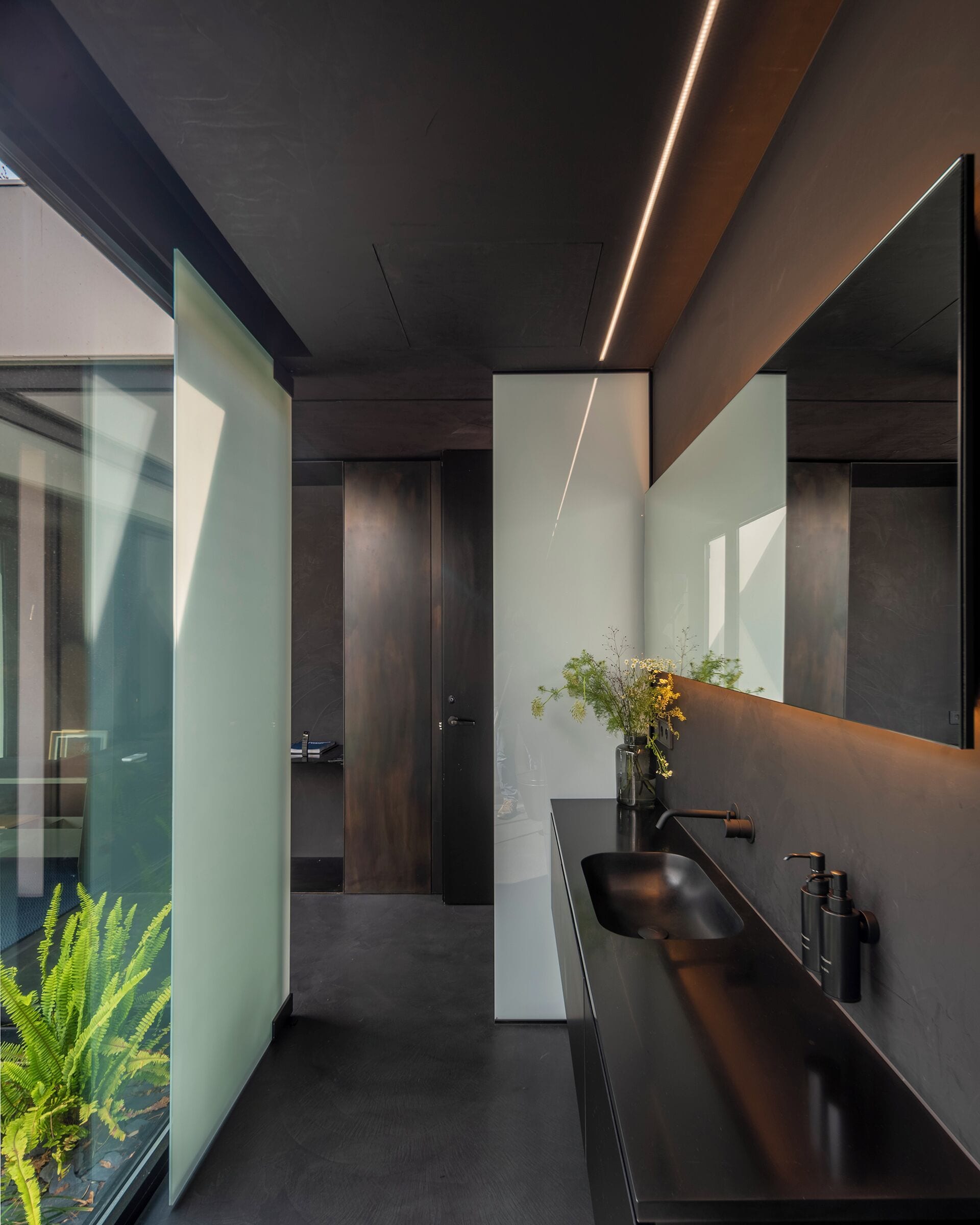 A modern bathroom with a long, dark sink, a large mirror, and a vase with flowers.
