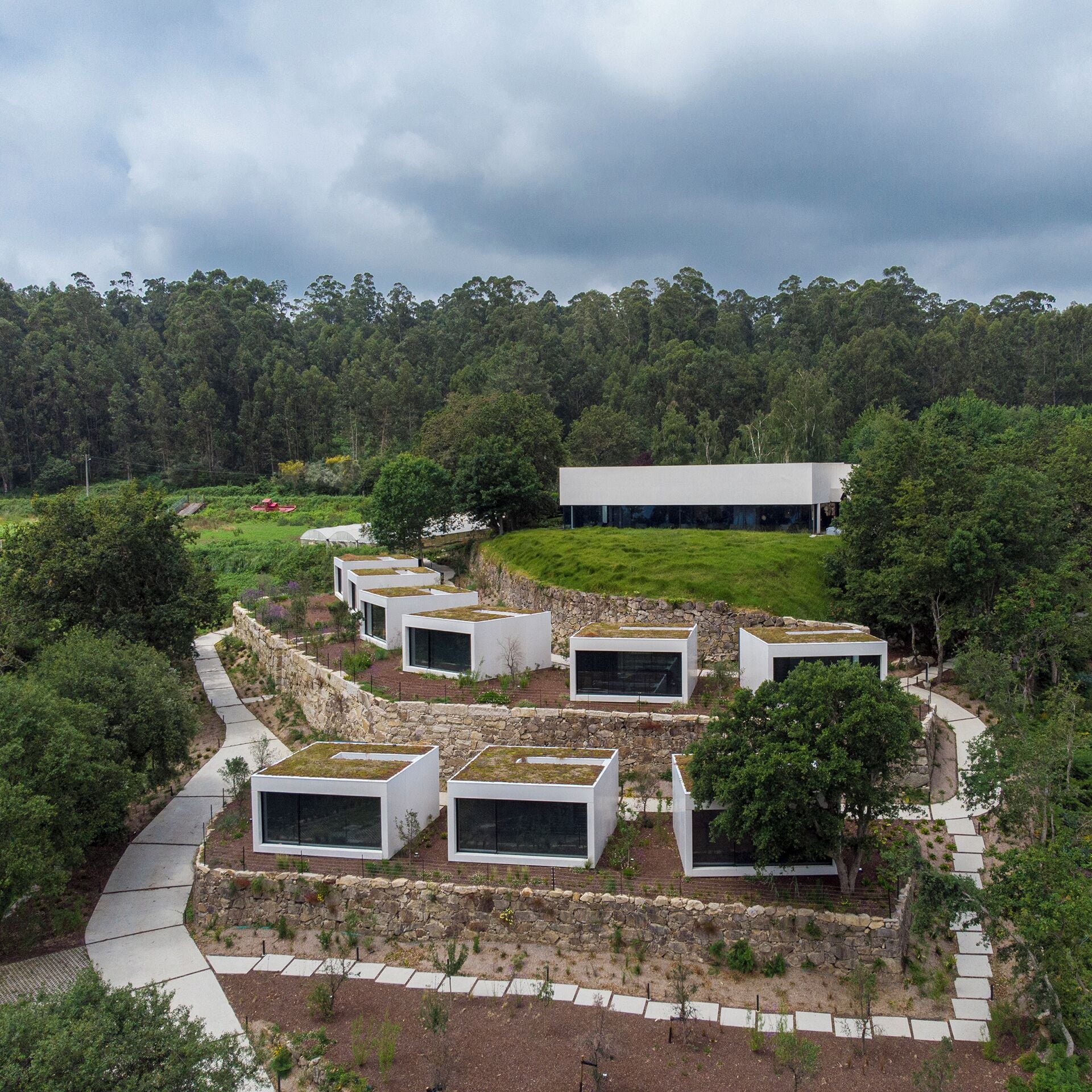 Un complejo moderno de edificios con varias estructuras blancas y techos planos, rodeado de exuberante vegetación y un paisaje boscoso.