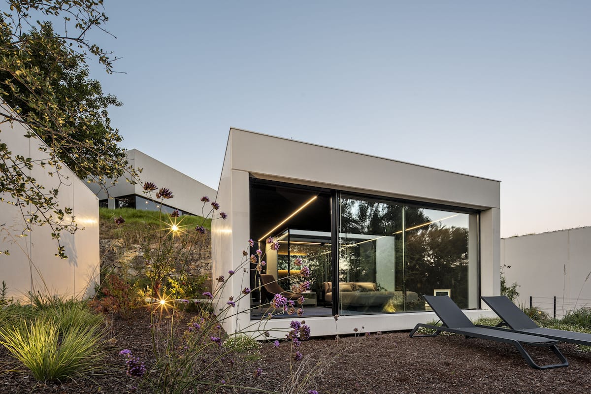 A modern house with a glass facade, a visible interior with a sofa and chairs, and a garden with plants and flowers.