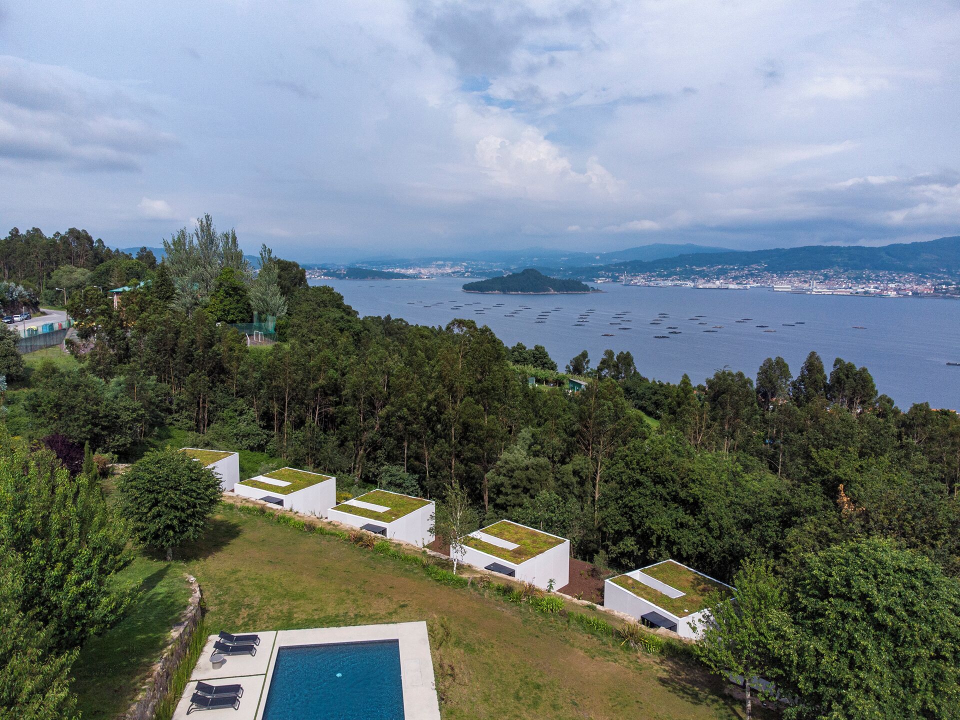 A modern resort with multiple buildings featuring flat roofs, surrounded by lush greenery and overlooking a large body of water with boats.
