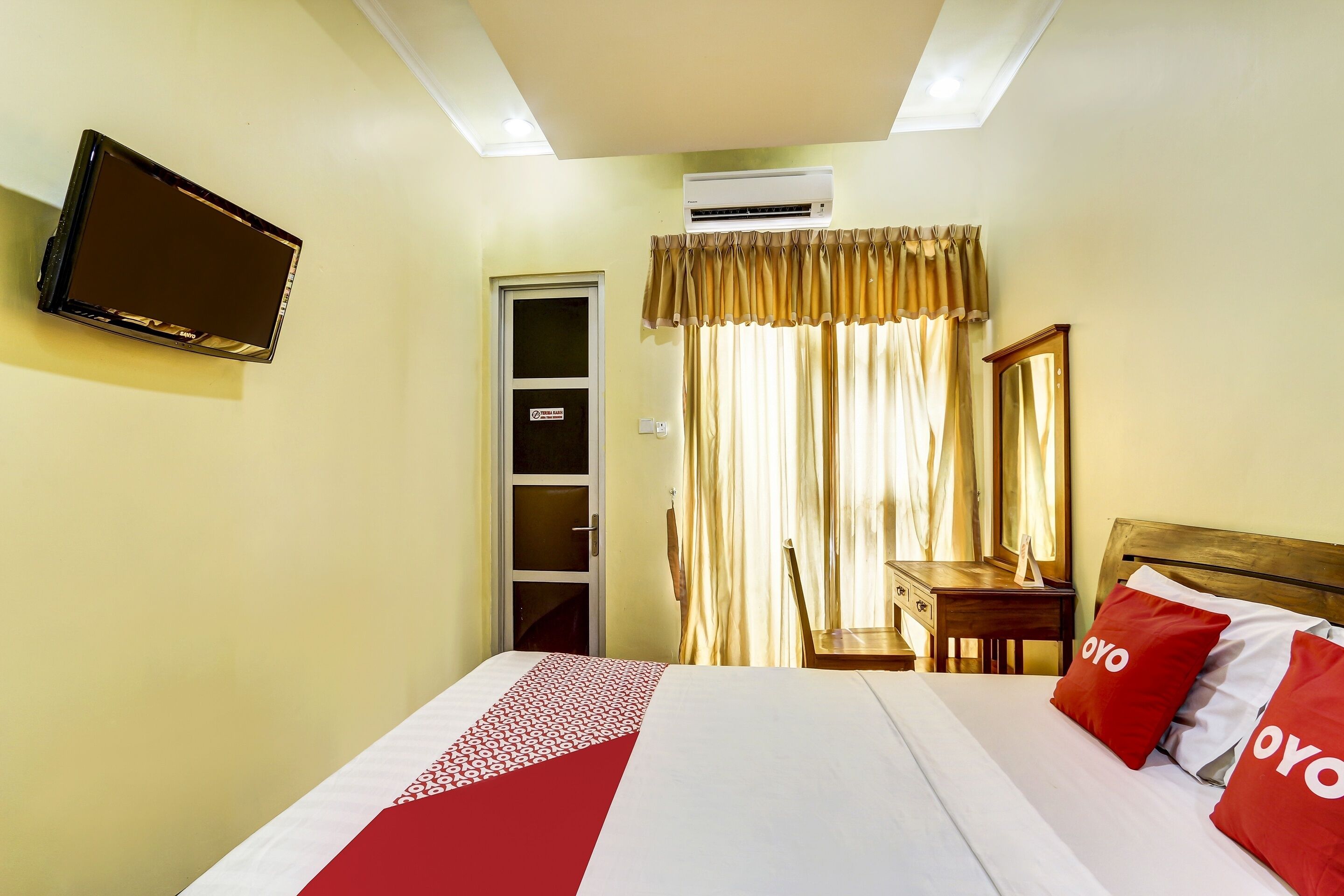 Une chambre d’hôtel avec un lit, un téléviseur fixé au mur, une fenêtre avec des rideaux et une table de chevet en bois.