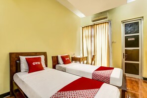 Habitación de hotel con dos camas, cada una con ropa blanca y almohadas rojas, un cabecero de madera, una mesita y una ventana con cortinas.