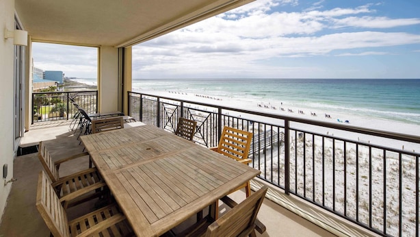 A balcony with a wooden table and chairs, overlooking a beach with a railing.