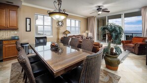 A spacious living area with a dining table, comfortable seating, a large potted plant, and a view of the ocean through large windows.