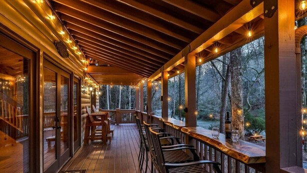 A covered outdoor deck with wooden railings, seating, and string lights. - Chardonnay River Chalet| Ellijay, GA (Ellijay)