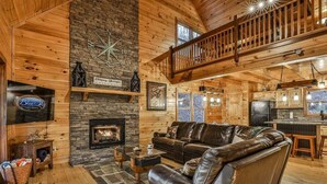 A log cabin interior with a stone fireplace, leather sofa, and a kitchen area. - Chardonnay River Chalet| Ellijay, GA (Ellijay)