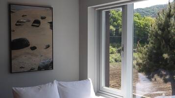 A room with a bed, a painting, and a window with a view of trees and hills.