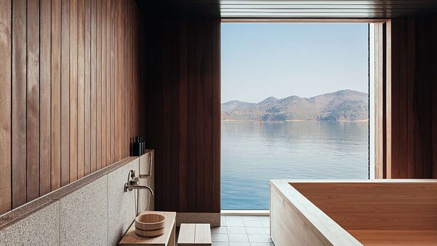 A modern bathroom with a wooden interior, a large bathtub, and a view of a lake and mountains. - IJE Namhae (Namhae)
