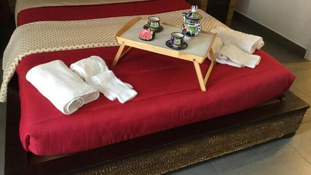 Una cama bien hecha con ropa de cama roja, una pequeña bandeja de madera con tazas de té y una tetera, y toallas enrolladas sobre la cama.