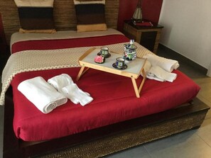 Una cama bien hecha con ropa de cama roja, una pequeña bandeja de madera con tazas de té y una tetera, y toallas enrolladas sobre la cama.