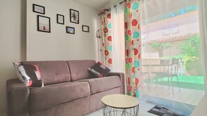 A living room with a brown sofa, a small round table, and a window with a view of a balcony and outdoor furniture. - Holiday home near the beach (Cap d'Agde)