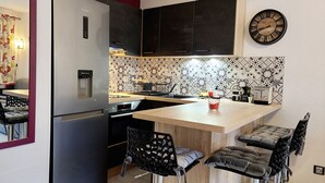 A compact kitchen with a refrigerator, a water dispenser, a microwave, a toaster, and a clock on the wall. There are bar stools and a patterned backsplash. - Holiday home near the beach (Cap d'Agde)