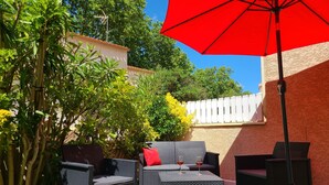 An outdoor patio area with wicker furniture, a red umbrella, and green artificial grass. - Holiday home near the beach (Cap d'Agde)